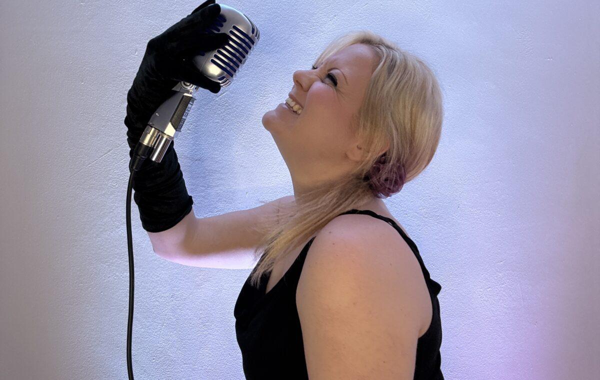 Tracy is a Wedding Singer Suffolk. She is laughing, looking up and holding a vintage microphone in the air.